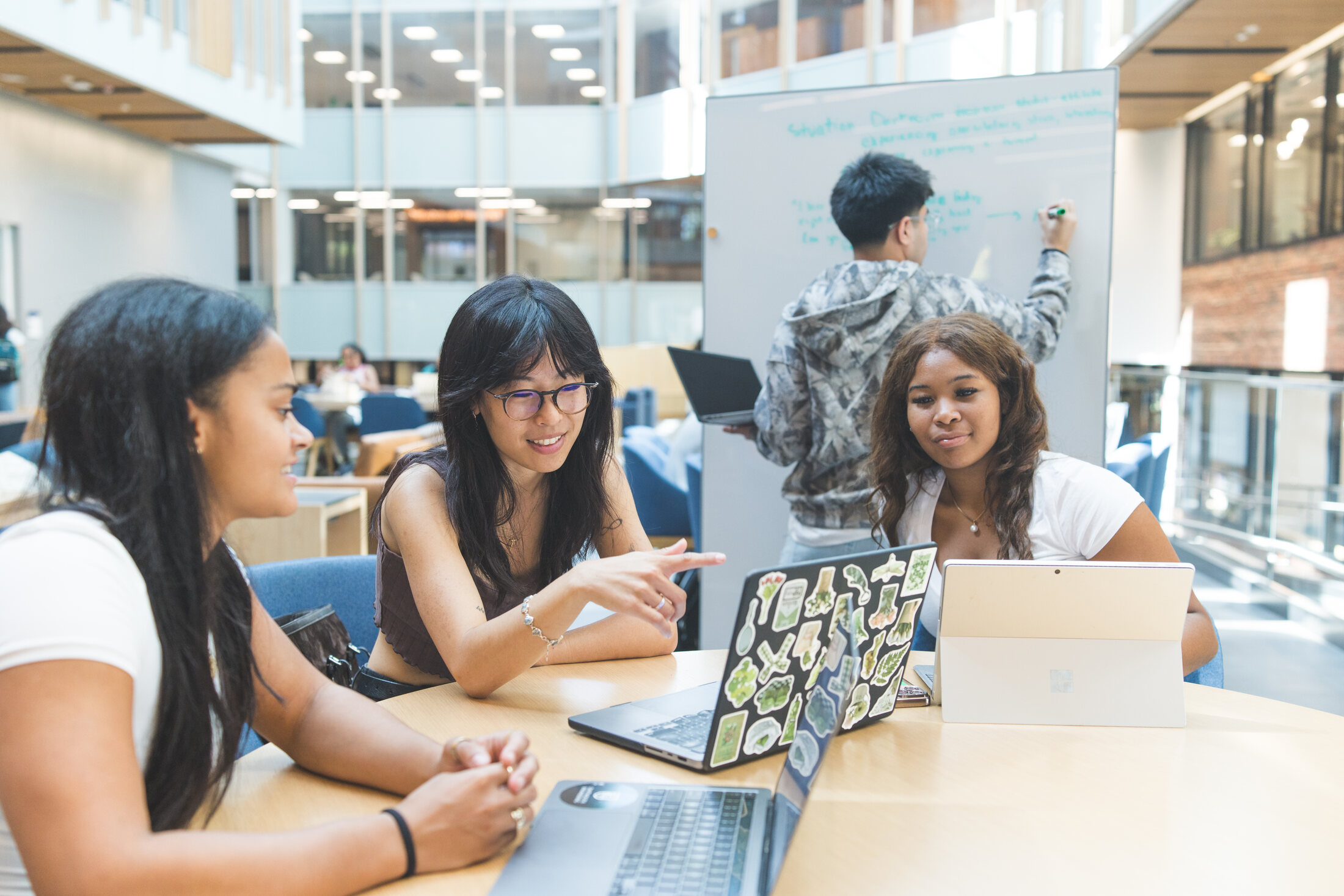 Three Evergreen students working together at a table while a teammate sketches on a whiteboard behind them